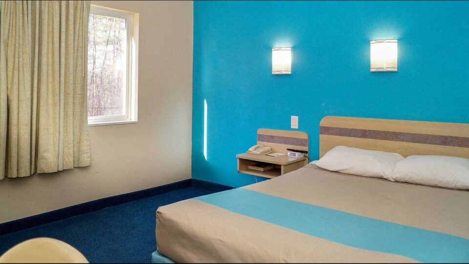 Image showing a simple hotel room with a bed with tan bedding and white pillows, wall behind bed is vibrant blue with white lamps.